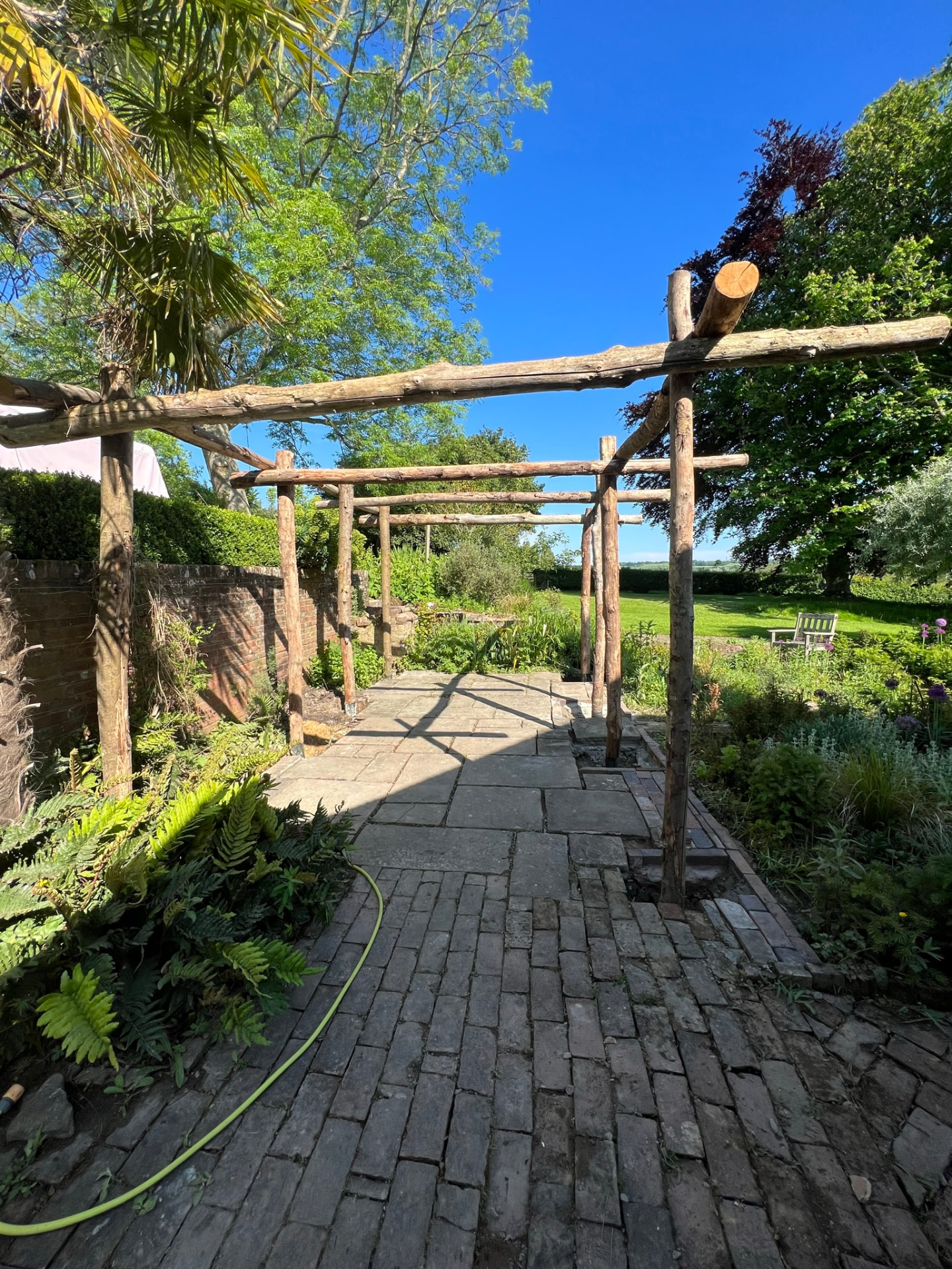 Rustic round-timber pergola running along a garden walkway of reclaimed brick and stone paving between clipped hedges and planted borders