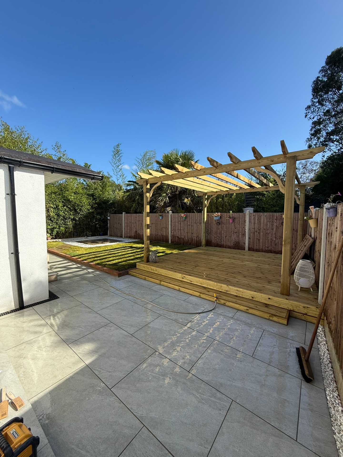 Completed garden with a large light grey porcelain tiled patio, a raised wooden deck with a new timber pergola and a lawn beyond a closeboard fence